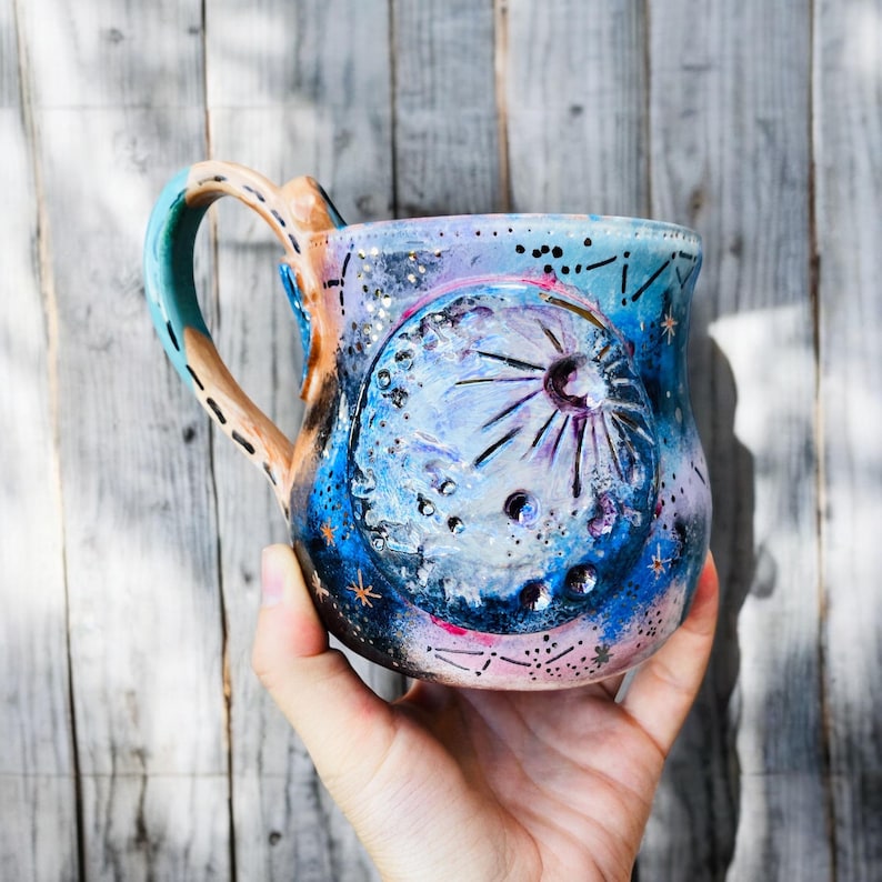 May include: A hand-painted ceramic mug with a galaxy design. The mug features a large moon with craters and stars, painted in shades of blue, pink, and white.