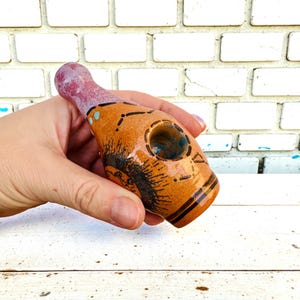 Op de afbeelding: Handgemaakte keramische pijp met een flesachtige vorm. De pijp heeft een bruin en oranje lichaam met zwarte en blauwe accenten. Het mondstuk is roze-paars. De pijp heeft een ronde kom en een decoratief ontwerp.