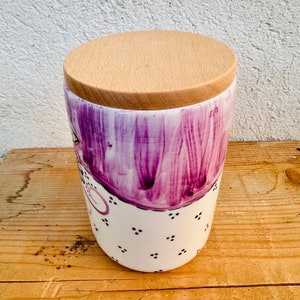 Puede incluir: Un recipiente cil&iacute;ndrico de cer&aacute;mica con un dise&ntilde;o de pinceladas moradas pintadas a mano en la parte superior y una base blanca decorada con peque&ntilde;os puntos negros. El recipiente est&aacute; cubierto por una tapa circular de madera clara.