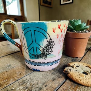 May include: A hand-painted ceramic mug with a peace sign and floral design. The mug features a blue peace symbol, red hearts, and a black floral outline. The handle is yellow and the base has a glittery blue band.