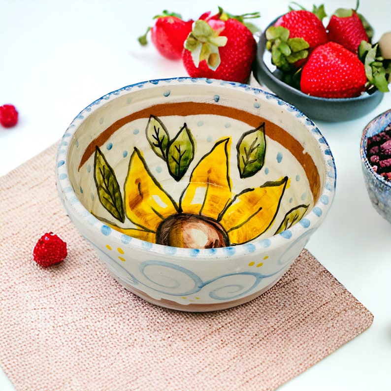 May include: A white ceramic bowl with a blue and yellow sunflower design. The bowl has a brown rim and is decorated with blue swirls and yellow dots.