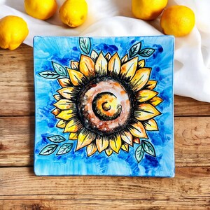 Puede incluir: Un plato cuadrado de cerámica azul con un diseño de girasol pintado a mano. El girasol es amarillo con un centro marrón y hojas verdes.