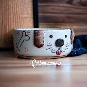 May include: White ceramic dog bowl with a brown and white dog face painted on the front. The bowl has a bone and red dots painted on the side. The bowl is on a wooden surface.
