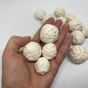 Può includere: Una mano che tiene una collezione di sfere di ceramica bianca con vari motivi in rilievo. Le sfere sono lisce e hanno una finitura opaca.