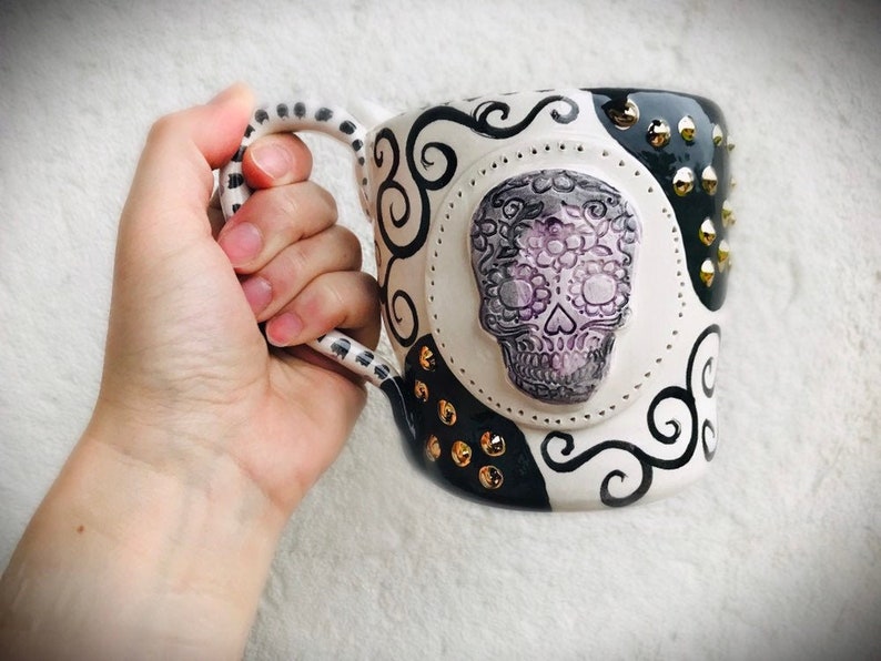 Pode incluir: Caneca de cer&acirc;mica branca com um design em preto e branco que apresenta uma caveira de a&ccedil;&uacute;car com detalhes em roxo e branco. A caneca tem uma al&ccedil;a preta com bolinhas brancas e tachas douradas ao redor da borda.