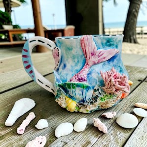 May include: A ceramic mug with a mermaid tail design. The mug has a blue and white background with a pink mermaid tail, coral, and seashells. The handle is white with blue and red accents. Seashells are scattered around the mug.