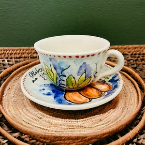 May include: A white ceramic teacup and saucer with a hand-painted design of leaves and pumpkins. The saucer has the text "Ottobre" written on it.