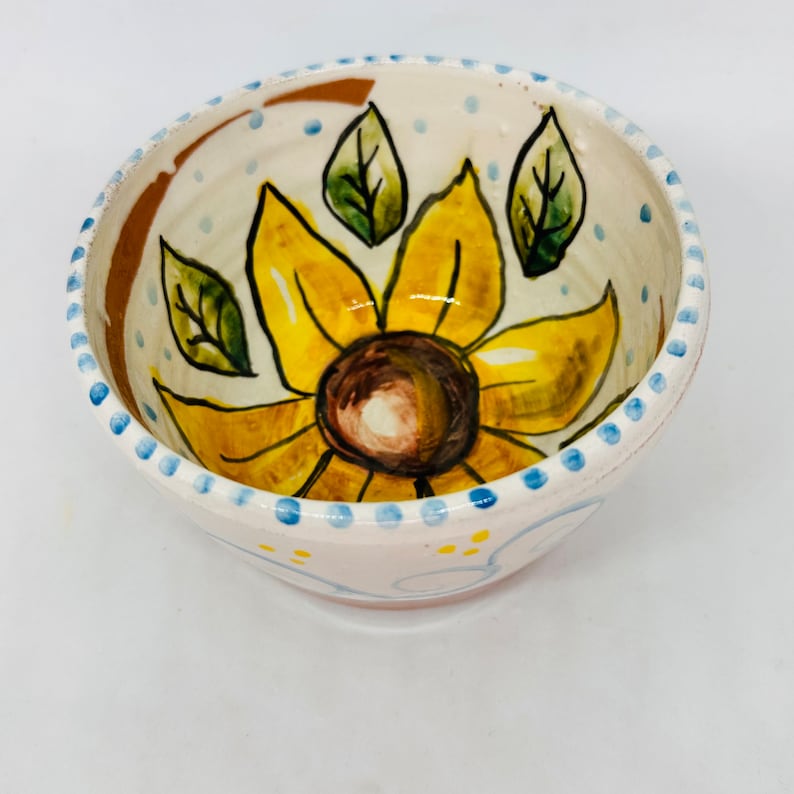 May include: A white ceramic bowl with a hand-painted sunflower design. The sunflower is yellow with a brown center and green leaves. The bowl has a blue and white dotted rim.