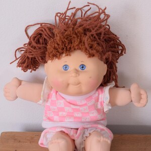 May include: A vintage doll with a soft, vinyl face, blue eyes, and reddish-brown yarn hair. The doll wears a pink and white checkered top and matching shorts. The doll is sitting on a wooden surface.