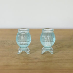 May include: A pair of small, light blue glass candle holders featuring a ribbed, rounded body and a flared top opening. Each holder is supported by three short, curved feet, creating a stable base for these translucent, decorative glass items.