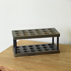 May include: A wooden test tube rack with two levels, each with multiple circular holes. The rack is stained a dark gray color and supported by four black posts. It sits on a wooden surface.