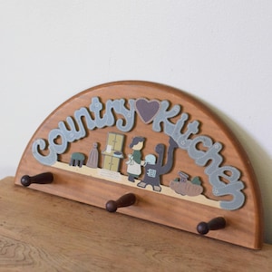 May include: A semi-circular wooden wall hook rack featuring three dark brown pegs. The top section displays the text "Country Kitchen" in grey script, accented by a small dark heart, and a decorative illustration of a person in a kitchen.