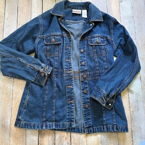 May include: A dark wash denim jacket with a button-up front, two chest pockets, and long sleeves. The jacket has a classic collar and a fitted waist. The brand name "Bill Blass" is visible on a tag.