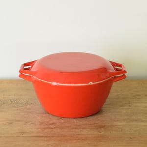 May include: A vibrant orange, lidded casserole dish. The round pot has two side handles and a matching lid. The dish sits on a wooden surface. A white accent line is visible between the lid and the pot.
