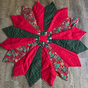 May include: A circular quilted tree skirt featuring a starburst pattern with alternating panels of solid red fabric, dark green fabric with a small cross print, and a multi-colored patchwork fabric with holiday motifs and Santa Claus figures.