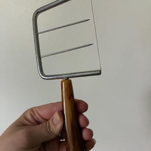 May include: A cheese slicer featuring a rectangular silver-toned metal frame with a thin wire cutting edge and two horizontal metal support bars. The handle is made of a smooth, polished brown material with a marbled, tortoiseshell-like pattern.