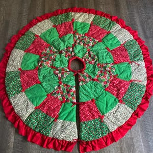 May include: A circular quilted tree skirt featuring a patchwork design of red, green, and cream fabric squares with various patterns. The skirt has a central opening, a slit for placement, and a ruffled red fabric border around the outer edge.