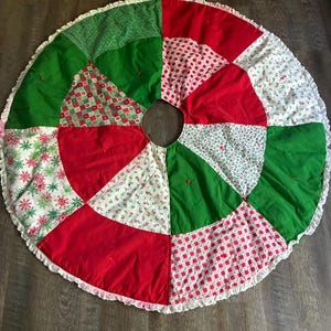 May include: A circular patchwork tree skirt featuring alternating triangular panels in red, solid green, and white with various patterns including red and green checkered prints, small floral motifs, and green snowflake designs, finished with a white ruffled edge.