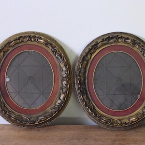 May include: A pair of matching oval picture frames featuring ornate, textured gold-toned outer borders and deep red inner matting. The center of each frame contains a glass pane with a subtle, etched geometric grid pattern of intersecting lines.