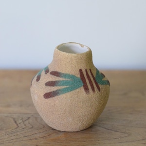May include: A small, round, tan-colored ceramic vase with a narrow neck. The vase is decorated with teal and maroon abstract designs. The interior of the vase is white. The vase is sitting on a wooden surface.