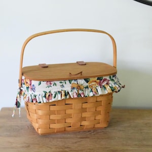 May include: A woven light brown wooden basket with a rectangular wooden lid and a single arched handle. The basket features a white fabric liner with a colorful floral pattern of roses and leaves, finished with a ruffled edge and a toggle closure.