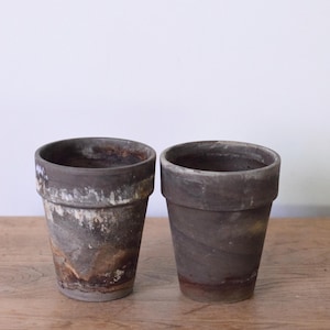 May include: Two cylindrical clay pots with a dark, weathered finish. The left pot displays mottled white and rust-colored mineral deposits, while the right pot features a smoother, dark charcoal surface with subtle brown tones near the base.