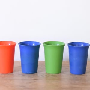 May include: A set of four drinking cups in vibrant solid colors, including one orange, two blue, and one green cup. Each cup features a smooth, tapered cylindrical shape with a slightly flared rim, standing upright in a single horizontal row.
