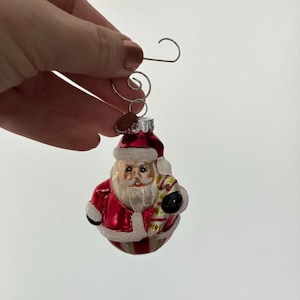 May include: A Christmas ornament shaped like Santa Claus, with a red coat, white trim, and a candy cane. The ornament has a silver hook for hanging.