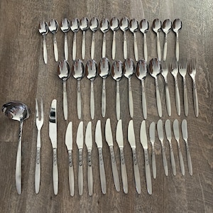 May include: A complete set of stainless steel silverware, including spoons, forks, knives, and butter knives, arranged on a wooden surface. The silverware has a simple, elegant design.