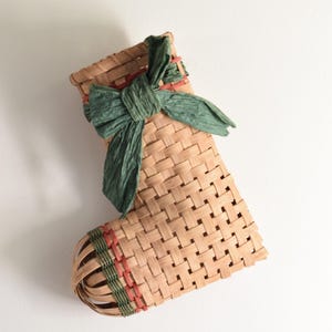 May include: A woven Christmas stocking made of natural-colored straw, with a green bow at the top. The stocking has red and green accents and is hanging on a white wall. A festive holiday decoration.