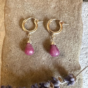 May include: A pair of gold hoop earrings with teardrop-shaped, pink gemstones. The earrings are displayed on a textured, light brown surface, with a sprig of lavender in the foreground. The earrings are a jewelry item.