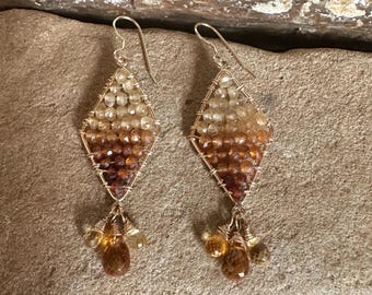 Puede incluir: Un par de pendientes de alambre dorado con un degradado de piedras preciosas de color marrón claro a oscuro. Los pendientes tienen forma de diamante y tienen un grupo colgante de tres piedras preciosas en forma de lágrima.