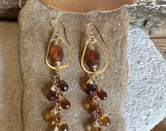 Puede incluir: Un par de pendientes colgantes dorados con un marco de alambre en forma de lágrima. Cada pendiente está adornado con un racimo de cuentas de piedras preciosas facetadas de color ámbar en tonos marrones, naranjas y amarillos.
