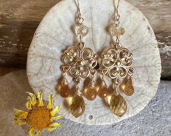 Puede incluir: Pendientes dorados con forma de araña con piedras preciosas de citrino. Los pendientes presentan un diseño de filigrana adornado con piedras de citrino en forma de lágrima colgantes. Una pequeña flor amarilla seca está en la esquina inferior izquierda.