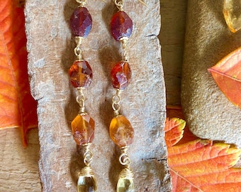 Puede incluir: Un par de aretes largos de oro con cuentas de piedras preciosas marrones y amarillas. Los aretes están colgando de una superficie de madera.
