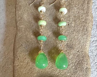 Pode incluir: Um par de brincos pendentes com uma disposição vertical de três pequenas contas redondas verdes e uma conta verde maior em forma de lágrima na base. As contas estão ligadas por um fio dourado e presas a ganchos dourados.
