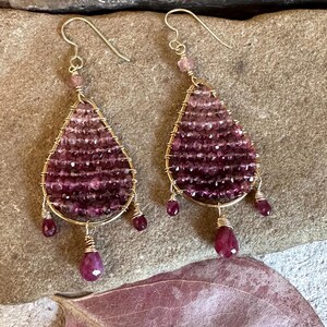 May include: A pair of teardrop-shaped earrings featuring gold wire frames filled with rows of faceted, deep magenta-colored gemstone beads. Each earring includes three dangling, pear-shaped gemstone drops at the base and a single bead on the hook.