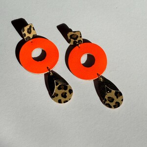 May include: A pair of neon orange and leopard print earrings. Each earring features a large orange circle with a smaller circle cut out of the center. The circle is attached to a smaller leopard print square and a teardrop-shaped leopard print dangle.