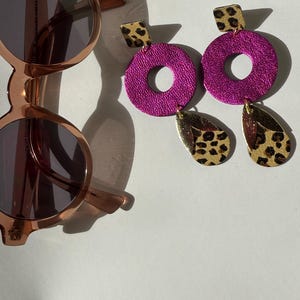May include: A pair of dangle earrings with a pink circle and a leopard print design. The earrings are made of leather and have a gold-tone finish.