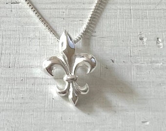 Puede incluir: Un collar de cadena de plata con un colgante de plata con forma de flor de lis. El collar está sobre un fondo de madera blanca.