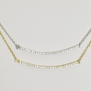 Op de afbeelding: Twee delicate kettingen met gebogen staven van sprankelende stenen. De ene ketting is goudkleurig, de andere zilverkleurig. Beide kettingen worden op een witte achtergrond gepresenteerd, wat hun elegante ontwerp en potentieel voor layering benadrukt.
