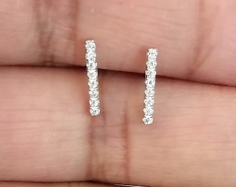 Puede incluir: Un par de pendientes pequeños de barra plateados. Cada pendiente es una línea recta de pequeñas piedras claras y brillantes. Los pendientes se muestran sobre un fondo claro.