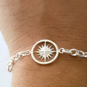 May include: A silver bracelet with a compass charm. The compass charm is a circle with a star in the center and a chain link bracelet.