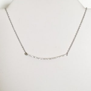 Op de afbeelding: Een zilverkleurige ketting met een gebogen hanger. De hanger is bezet met kleine, heldere kristallen. De ketting wordt gepresenteerd op een witte mannequin buste.
