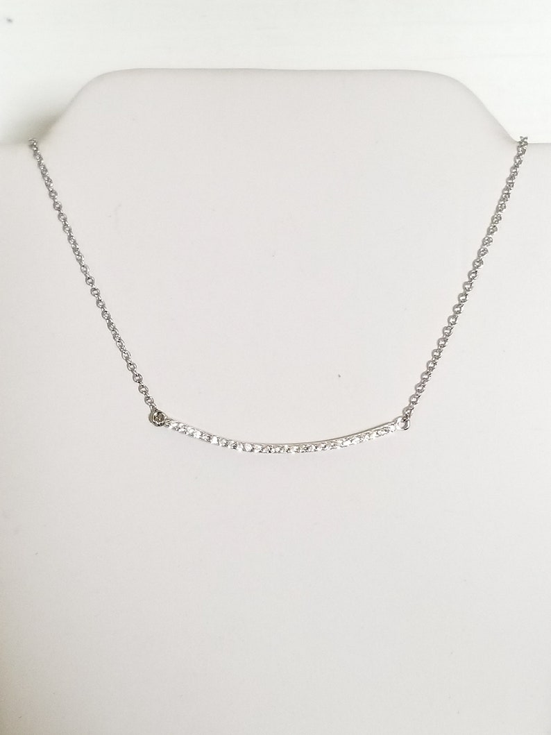 Op de afbeelding: Een zilverkleurige ketting met een gebogen hanger. De hanger is bezet met kleine, heldere kristallen. De ketting wordt gepresenteerd op een witte mannequin buste.