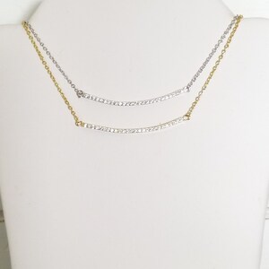 Op de afbeelding: Twee delicate kettingen, gepresenteerd op een witte mannequin. De bovenste ketting is zilverkleurig, de onderste goudkleurig. Beide hebben een gebogen hanger met kleine, sprankelende steentjes. De kettingen zijn fijn en elegant.
