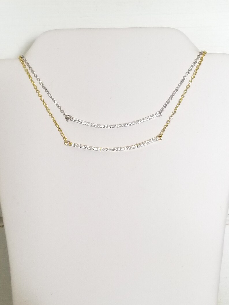 Op de afbeelding: Twee delicate kettingen, gepresenteerd op een witte mannequin. De bovenste ketting is zilverkleurig, de onderste goudkleurig. Beide hebben een gebogen hanger met kleine, sprankelende steentjes. De kettingen zijn fijn en elegant.
