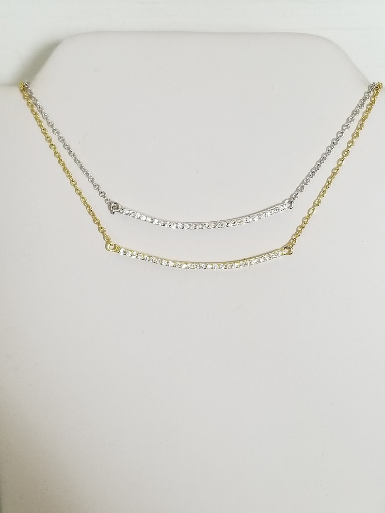 Op de afbeelding: Twee delicate kettingen met gebogen staven van sprankelende stenen. De ene ketting is goudkleurig, de andere zilverkleurig. Beide kettingen worden op een witte achtergrond gepresenteerd, wat hun elegante ontwerp en potentieel voor layering benadrukt.