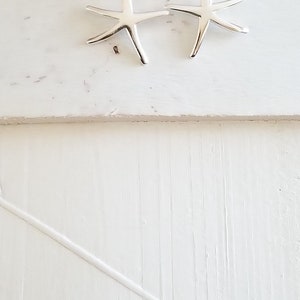May include: A pair of silver starfish earrings hanging from a piece of twine against a white wooden background.