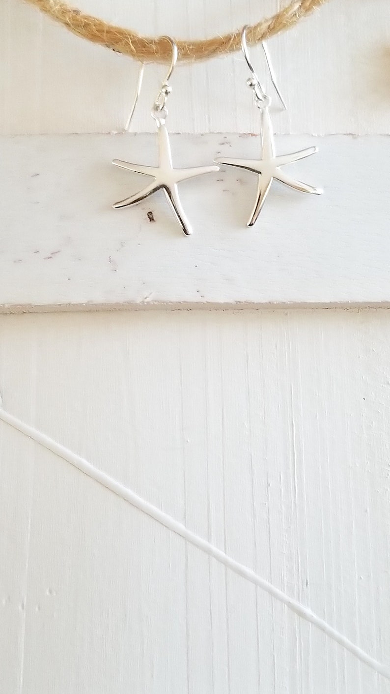 May include: A pair of silver starfish earrings hanging from a piece of twine against a white wooden background.
